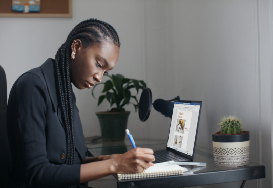 Women working from home holding a pen and writing on a note book in front of a laptop.