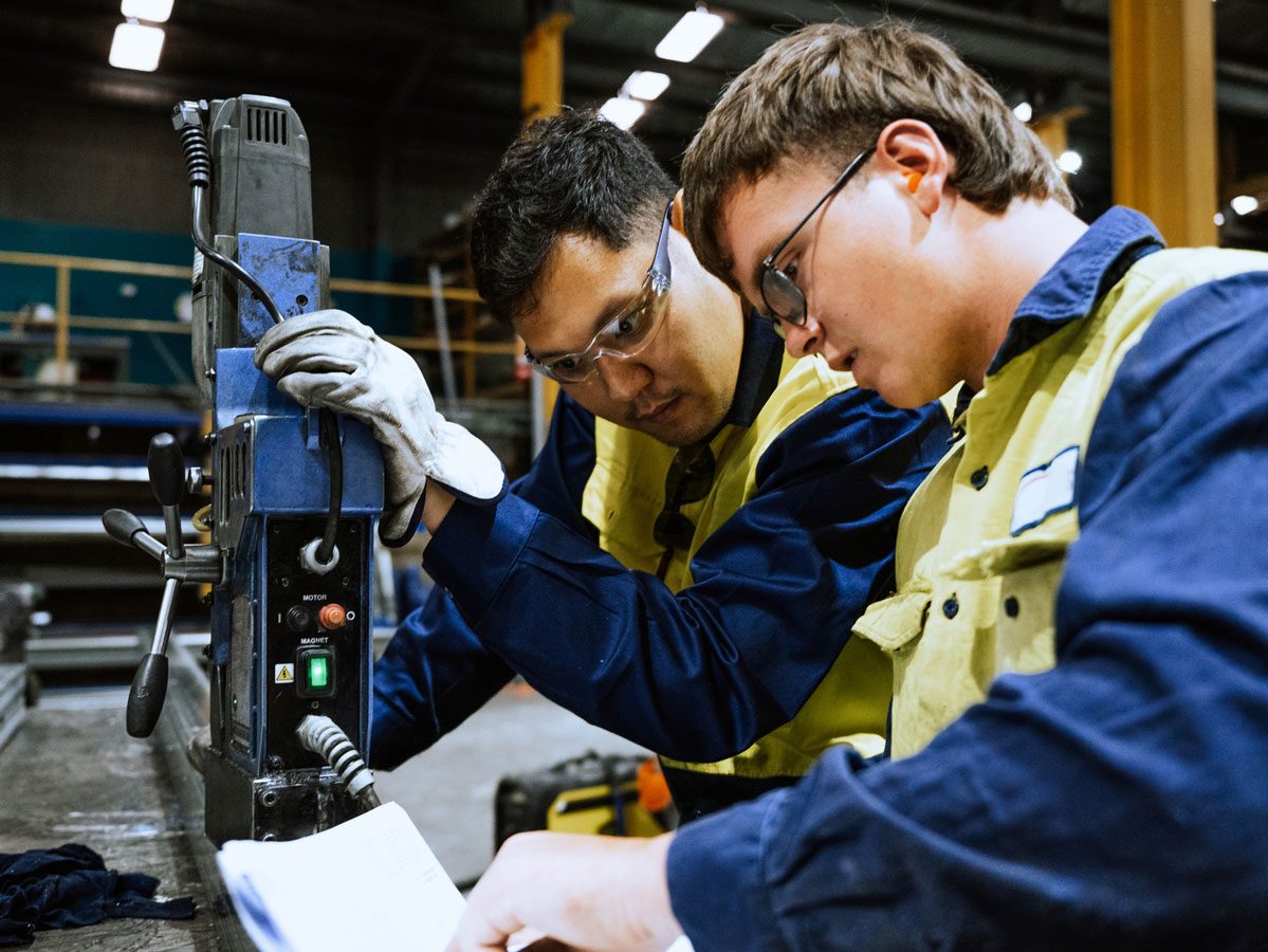 Alt: Industrial workers in safety gear collaborating over a machine, demonstrating practical skill application and hands-on learning.