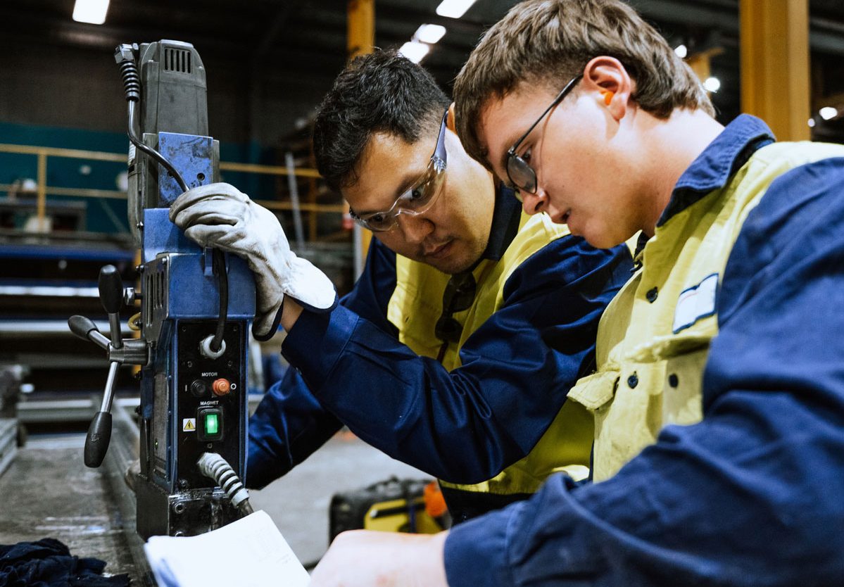 Alt: Industrial workers in safety gear collaborating over a machine, demonstrating practical skill application and hands-on learning.